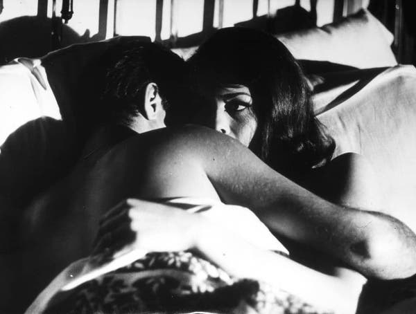 This image depicts a romantic scene with two people lying close together on a bed. They are partially covered by sheets, and the man's arms are wrapped around the woman. The woman has her eyes closed, while the man is looking at her, their faces expressing intimacy. There is no text visible in this image. The style of the image suggests it might be from a film or a TV show, given the cinematic quality and composition.