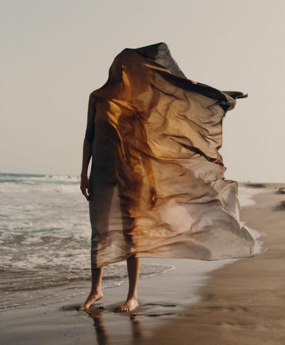 Photograph. A color photo of a woman wrapped in a scarf on the beach. A young woman stands on a beige sandy beach with white sea waves, completely wrapped in a semi-transparent cloth. Her head is also covered. The wind plays with the cloth, drawing folds and blowing it next to her body. The light is orange and creates a warm vacation atmosphere