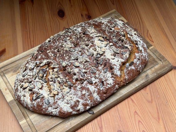 A photo of a big loaf of bread on top of a wooden cutting board. It has flour and nuts on top. 