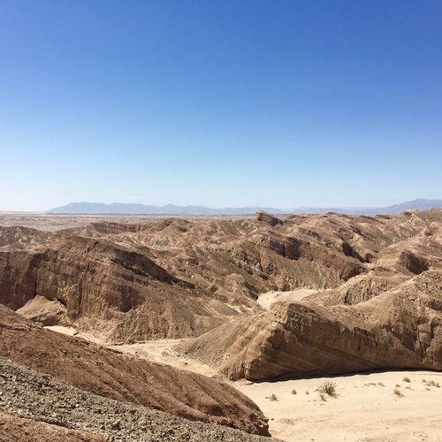 iPhone photo of the Anza-Borrego badlands with massive striated rocky terrain & canyons in the foreground & blue mountains & a hot blue summer sky in the background