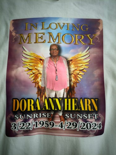 Photo of the front of a t-shirt. On the shirt is a photo of an older black lady with black glasses and golden wings. Above her is the text "IN LOVING MEMORY". Below her is three lines of text: "DORA ANN HEARN", "SUNRISE SUNSET", "3/22/1959-4/29/2024"