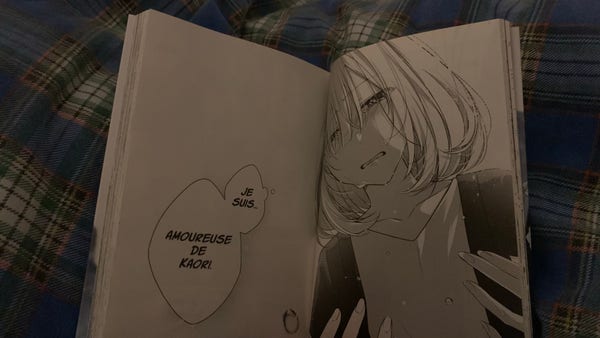 Photo d’un manga entrouvert qui se nomme « notre été éphémère ».
Il est posé sur une couette à motifs écossais, de couleur dominante bleue.
Sur la page de droite, le visage et le buste d’une jeune femme aux cheveux clairs mi-courts, elle est en pleurs.
Sur la page de gauche, apparait ses pensées où elle se dit : « Je suis amoureuse de Kaori ».