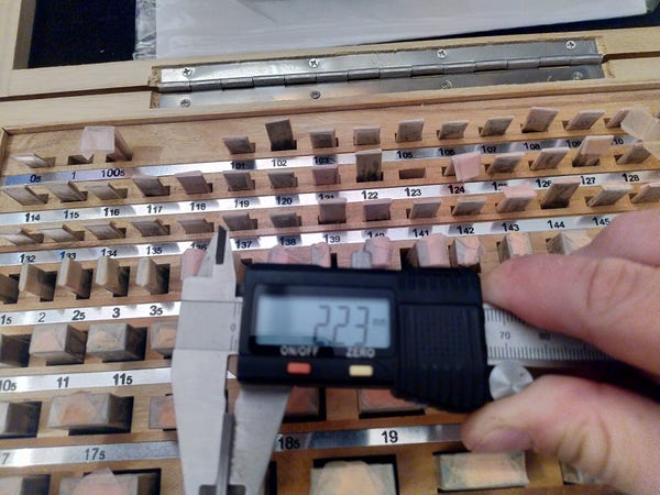 A Vernier caliper holding a pair of gauge blocks. The caliper shows 2.23mm. Behind the caliper a set of gauge blocks shows 1.00mm and 1.23mm missing. 