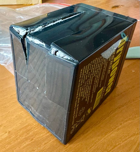 A black roughly-cube lead-acid battery, which has split open in multiple places