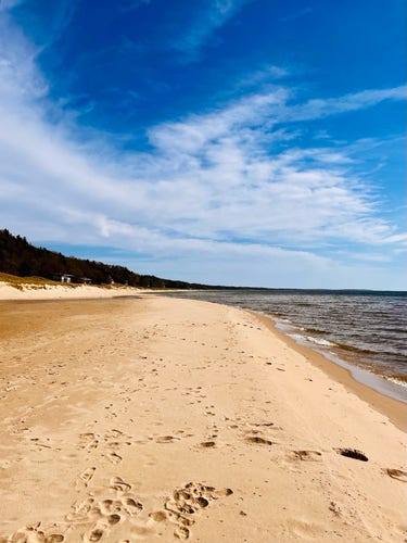 A sandy beach with footprints, bordering a calm Lake Michigan, under a blue sky with wispy clouds. Trees line the beach in the distance.