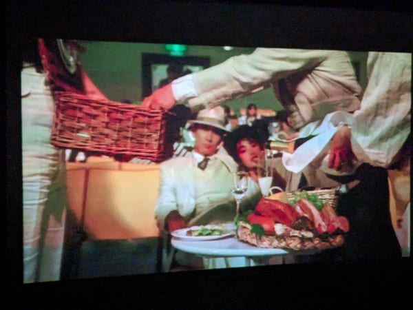 Still from movie with champagne being poured at a table set up at the front of a movie theatre