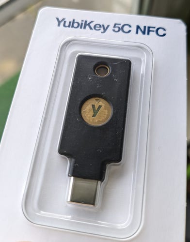 A YubiKey 5C NFC in its original blister-and-cardboard packaging with no visible exterior damage. However, the key has, visible through the blister plastic, obvious fingerprint remains on their touch surface (the gold one with the Yubico logo).