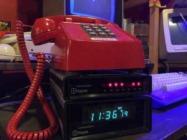 A red telephone sitting on top of two Hayes Stack devices: a SmartModem 300 baud modem and a Chronograph. A 1702 monitor, C64c, and a DEC VT-220 is in the background. It’s an older picture so the day of week and time are wrong. 