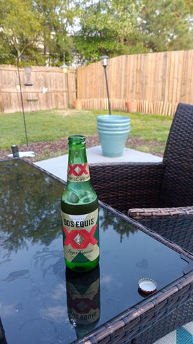A green bottle of Dos Equis with a high end wicker chair, a mirrored black table, and some plant pots and grass in the bacground