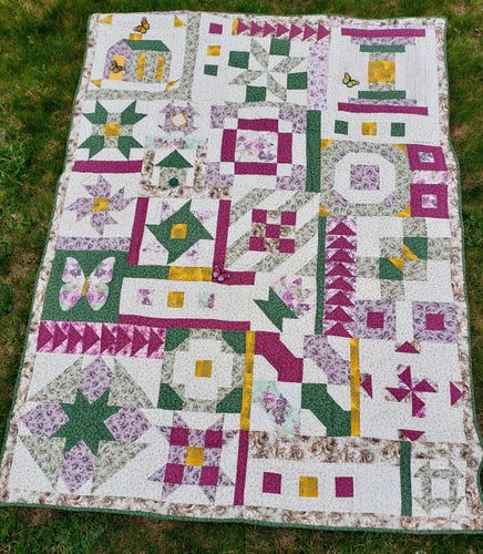 a  photo of a quilt on grass, it is a whimsical scene, a house and a butterfly and other geometric abstract designs, done in purples and greens and burgundy and gold on a white background with faint floral print 