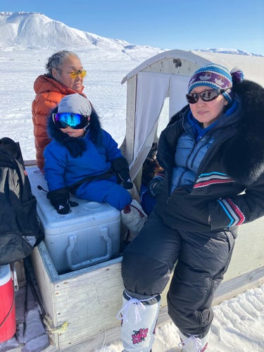Three people sit at the back of an iglutak (shelter) on a qamutiq (sled). The three are Inuit, an elder woman in an orange parka, a young boy in a royal blue snowsuit and reflective goggles. And a young woman in a black parka with layers underneath. The landscape and sea ice are covered in snow.  A mountain and bright blue skies fill the background. 