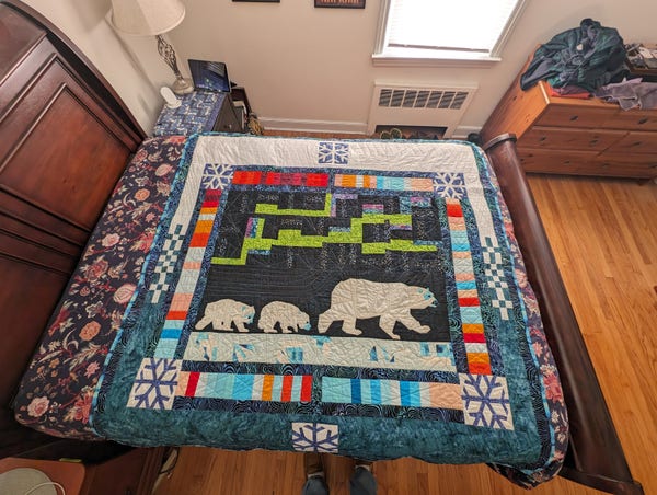 A quilt spread on a bed. The center panel shows 3 polar bears, an adult and 2 cubs, walking across ice, a blocky aurora is above them. Around the panel is a striped border with colors ranging from dark blue to dark red with blue and white snowflakes at each corner. The top is white with another snowflake block in the middle. The bottom is a rich sea green with a sixth snowflake block.