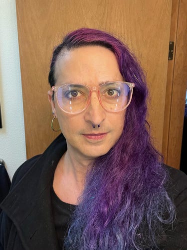 Selfie of Nora standing in front of a wooden door. Shes a middle-aged white trans woman looking at the camera with a slight smirk on her face. She’s wearing large pink-framed glasses, hoop earrings, and has long purple hair all combed over to one side, with flecks of silver glittery fairy hair in it. The sides of her head are shaved. 