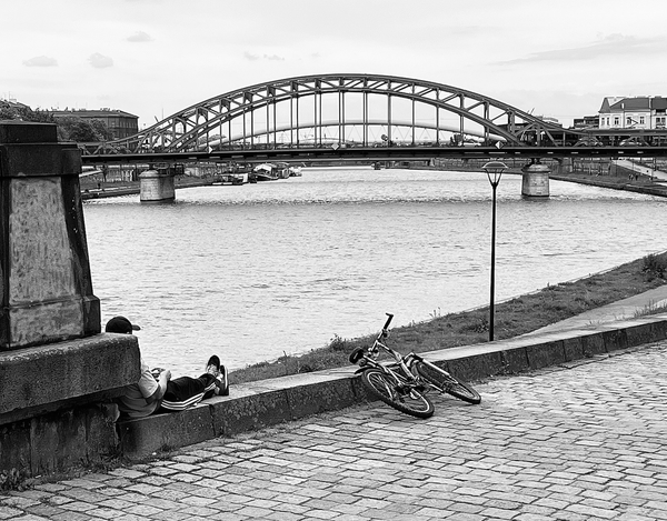 Black and white photography showing a section of the Vistula River in the city of Krakow with its two banks. An old cobbled road leads to the right bank of the river, at the beginning of which stands a stone pillar. Behind the pillar can be seen the figure of a seated young boy staring at his phone screen. Just in front of him lies the bicycle on which he arrived. The horizon is closed by an old steel truss bridge connecting the two banks of the river (the city's two districts). Behind the bridge, several boats are moored on the left.