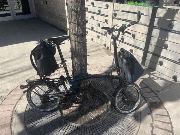 A black Brompton e-folding bike with a collapsed “shopping cart” bag mounted on the front block and a briefcase-style bag mounted on the SeatPostHugger. It’s unfolded and leaning against a tree. 