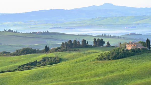 The rising sun reveals a Tuscan farmhouse amidst rolling fields of green spring wheat.