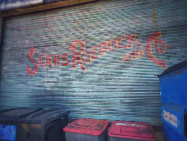 Old industrial rolling garage door with "Sears and Roebuck's" on it with garbage cans and a dumpster in front of it.