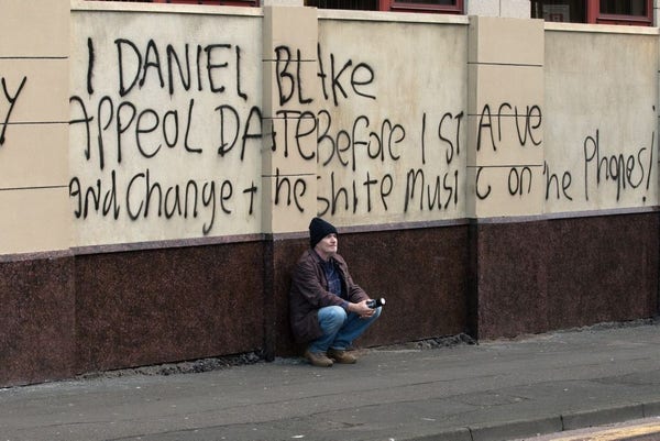 Un hombre en cuclillas delante de una pintada en una facha que dice: Yo Daniel Blake exijo una fecha para mi apelación antes de morir de hambre y cambiad la música de mierda del teléfono 