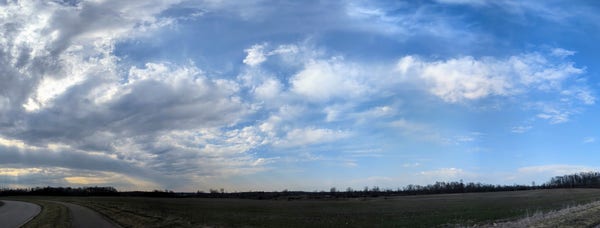 A panoramic view of a barren field in springtime with clouds to the left and sunlight and rays of sunlight showing through it in blue skies and slightly Cloudy Skies to the left