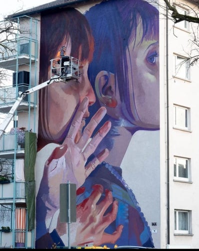 Streetartwall. A new mural is currently being created on the outside wall of a four-storey modern building. A man is standing on a lifting platform on the white wall of the building. Next to him are two painted female heads in purple and blue. The artist during the creation process. 21/03/2024