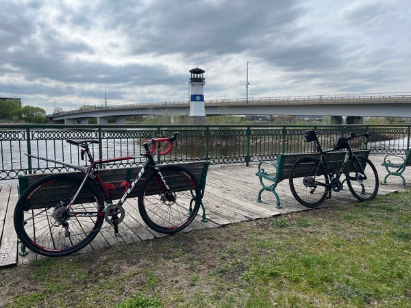 The image shows two bicycles parked on a wooden boardwalk overlooking a body of water. Both bicycles are gravel bikes.  The bike on the left has matte black frame with crimson red and brilliant white accents. The bike on the left is British racing green, with bright lettering.

In the background, there is a bridge spanning the water. In front of the bridge is a white light house.

The sky is overcast with greyish-blue clouds.

In the foreground, the boardwalk is preceded by a grassy area. A few benches are also visible along the path.
