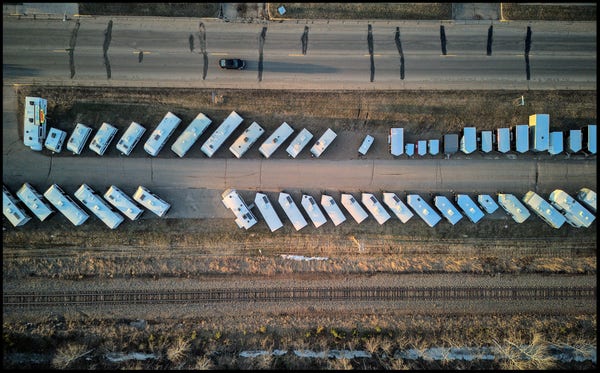 Looking straight down at a parking lot with two long rows of camper trailers for sale. There is a road above the lot and a railroad track is running underneath the lot.