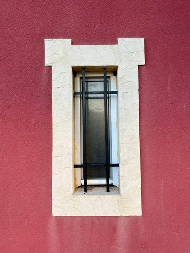 A narrow window with frosted glass and iron bars, set in a broad frame of white stone on a red wall