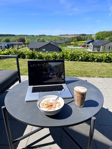 Outdoor setting. Blue sky and fields in the background. In the foreground is a patio with a table. In the table is a MacBook showing a cycling race along with a bowl of milk and cookies and a caffe latte.