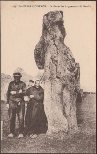A vintage postcard of c.1920 showing an elderly couple standing beside the "Géant des Alignements du Moulin" at St-Pierre-Quiberon on the Quiberon Peninsula in Brittany. The prehistoric stone is about twice the height of the old man and, behind, several rows of standing stones lead off into the distance.
