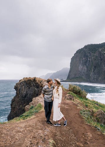 A couple standing on the edge of a cliff, with the ocean and cliffs in the background
