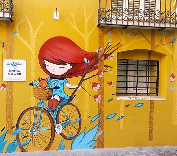 Streetartwall. On the outside wall of a yellow-painted house in Valencia is a charming little mural of a manga-style cyclist. A little girl with long red hair, a turquoise dress and red shoes is riding a turquoise bicycle through green grass. Her hair is blowing in the wind.  She has her little brown cat in a basket in front of her and is holding a branch with little laughing houses hanging from strings. Green leaves fly over the wall. Behind a real barred window "hangs" a single little painted house attached to a string. (On the wall of the house hangs a sign for a bicycle rental shop)