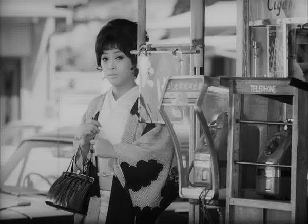 A woman is close to a telephone booth. She is holding a dark purse. Her hair is dark and straight. She is wearing a kimono-like coat. There is a car behind her.