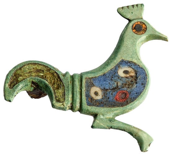 The picture shows a fibula (brooch) in the shape of a peacock. It's made of bronze and was inlaid with coloured enamel. This is broken out in section of the tail. The body is blue with a white and red circle pattern. The eye is orange with a black pupil.
