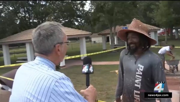 A reporter talks to a man covered in paint flecks and wearing a shirt that says “Paint Life”