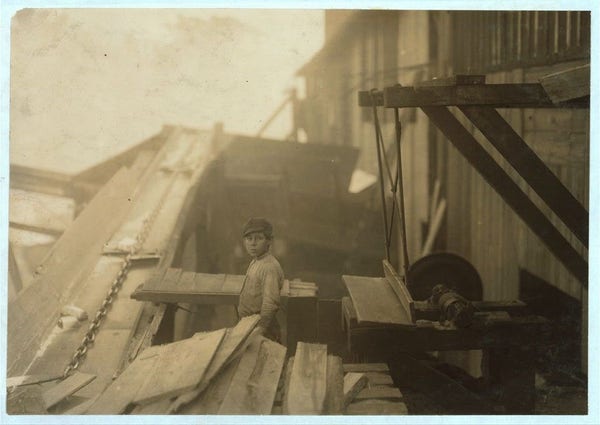  The image is a black and white photograph depicting an industrial setting. There is a person standing amidst various pieces of what appears to be metal or wood, suggesting some form of dismantling or construction activity. The individual is wearing attire that suggests manual labor, possibly work clothes. In the background, there is a structure with a sign that reads "Miller & Vidor Lumber Company."

The text overlay describes the context and details about the subject of the photograph: Charlie McBride, who is described as a twelve-year-old boy working at this lumber company. His job involves handling slabs out of a chute and passing them onto another person for sawing on an unguarded circular saw. The text also notes that Charlie works for four bits (twenty-five cents) per day, often for ten hours, and that the company is located outside Beaumont, Texas. It highlights the risks associated with this type of work, including exposure to weather without a roof, as well as the inherent hazards in handling machinery.

This image serves as a historical representation of child labor practices in the early 20th century, particularly in industries like lumber where such practices were common despite the risks involved.