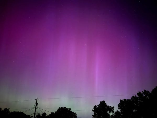 An aurora borealis. Pinkish hues with some vertical streaks. Trees and power lines are at the bottom of the frame. 