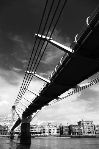 Black and white shot of the modern Millennium Bridge seen from the bottom left, from the banks of the Thames. The harsh light makes the underside of the bridge appear splendidly deep black, running diagonally through the picture and dominating the entire shot. A few snow-white, dramatic cloud formations glide across the otherwise dark sky. The city can be seen in the background on the other bank; St Paul's Cathedral is on the far left.