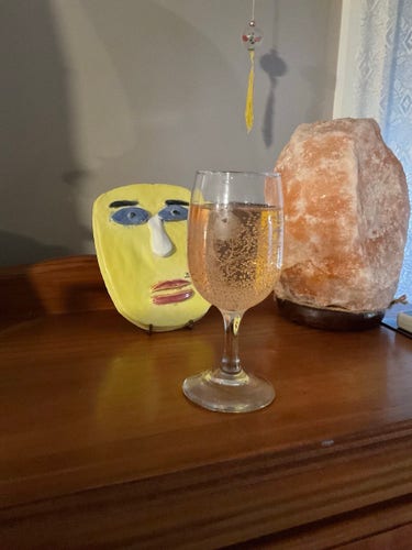 Glass of pale pink bubbly Rosé on top of a wooden cabinet. A yellow ceramic face with blue eyes, a white nose & red lips is to the left of the glass & a pink salt lamp is on the right. There is a tiny hanging ornament above the glass of wine. 