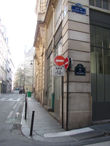 Ein urbanes Bild in Paris: eine leere Straße mit Häuserfassade und einer Laterne im Stil des 19. Jahrhunderts. In Bildmitte eine Hausecke mit roten Durchfahrverbotsschildern, darunter ein eckiges weißes Schild mit "SAUF" und einem Fahrrad-Piktogramm. Ein blaues Straßenschild an der Hauswand weist auf die Rue de la Ville Neuve, 2e Arr hin. Im Bildhintergrund gleißendes Sonnenlicht, ein parkendes Auto am Ende der Straße, ein abgestelltes Fahrrad, eine grüne Mülltonne. DIe Fahrradmarkierung auf der Straße ist sehr schmal.