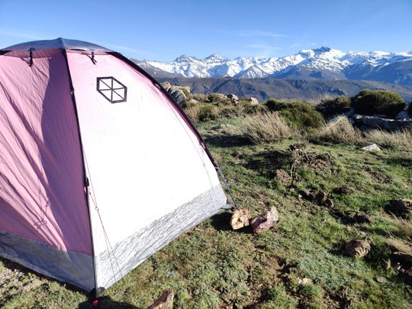 A pink tent with a Samaya logo on lies on a flat grass surface. Beyond are snow covered mountains