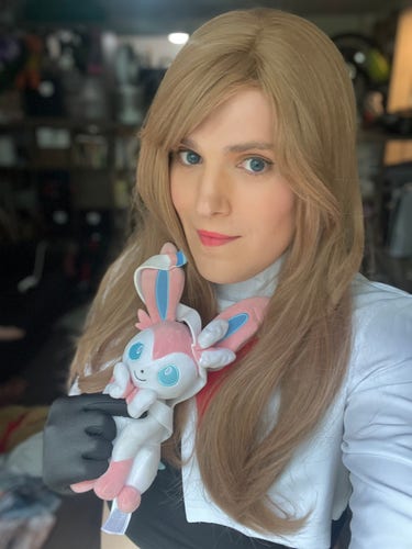 A trans woman with blue eyes and blonde hair holds a slyveon (the eevee evolution with trans flag colors) that she stole from some twerp. She is dressed a member of team rocket.