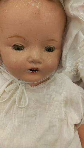 An antique baby doll with light skin, empty dark eyes hidden by lashes. It wears a white, old fashioned nightgown and a baby bonnet that ties under it's chin. The paint is chipped on the cheeks and forehead.