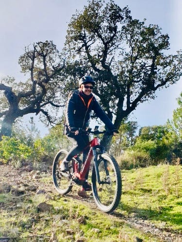 White middle-aged women (me) riding a red fully-suspended electric mountain bike in a natural setting with trees and grass on a sunny day. I wear sun glasses and a grey helmet. I’m smiling. 