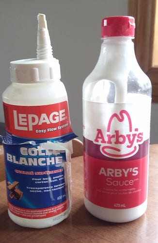 Colour photo of two plastic bottles. The one on the left held LePage white wood glue and was about half full before being needed today. It cracked on the right side around where the label and bottle was subsequently cut with scissors, so I could transfer the contents to the bottle beside it -- an empty and clean clear Arby's Sauce bottle, that has a red flip cap, so will function well the next time some of the remaining glue may be needed.