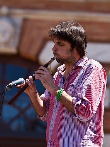 Un joueur de Duduk, vêtu d’une chemise à rayures rouges et blanches, joue de cet instrument traditionnel arménien en plein air. Il porte un bracelet vert au poignet et est proche d’un microphone pour amplifier le son mélodieux du Duduk.