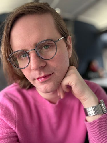 Photo of mb, a white transfeminine nonbinary person in a bright pink sweater, sparkly pink lip glosses and glasses
