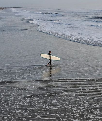 From high up on the Jacksonville Beach Pier, looking down at the wet beach in low tide, a lone surfer in tight black rubber wet suit, carrying a surf board under one arm walks out into the waters as the waves splash along the coast.