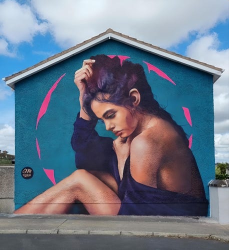 Streetartwall. The charming mural of a young woman was sprayed/painted on the outside wall of a single-storey detached house. The background is blue-turquoise with some pink graffiti arches forming a circular frame around the woman. She is depicted from the side, wrapped in a black cloth and sitting half-naked on the floor. She wears her long brown hair in a plait and grasps the hair on her head with one hand. The other hand is under her chin. She looks down, lost in thought. The artist's signature Graffmatt can be seen in a small black field on the left. In the photo, only one street can be seen in front of the mural. The house is a corner house.