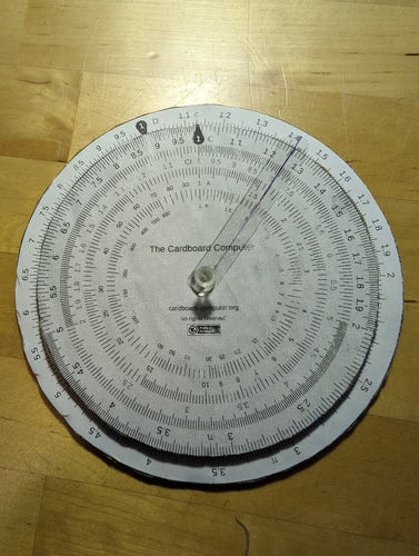 A circular slide rule printed out on paper and glued to thick cardboard backing, with a thumbtack as a pivot.