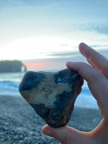 A hand holds a vaguely heart-shaped, multicolored beach pebble, bluish gray, light brown, yellowish white… The backdrop is the rocky beach, the sea, and the Étretat cliff and needle in the sunset.